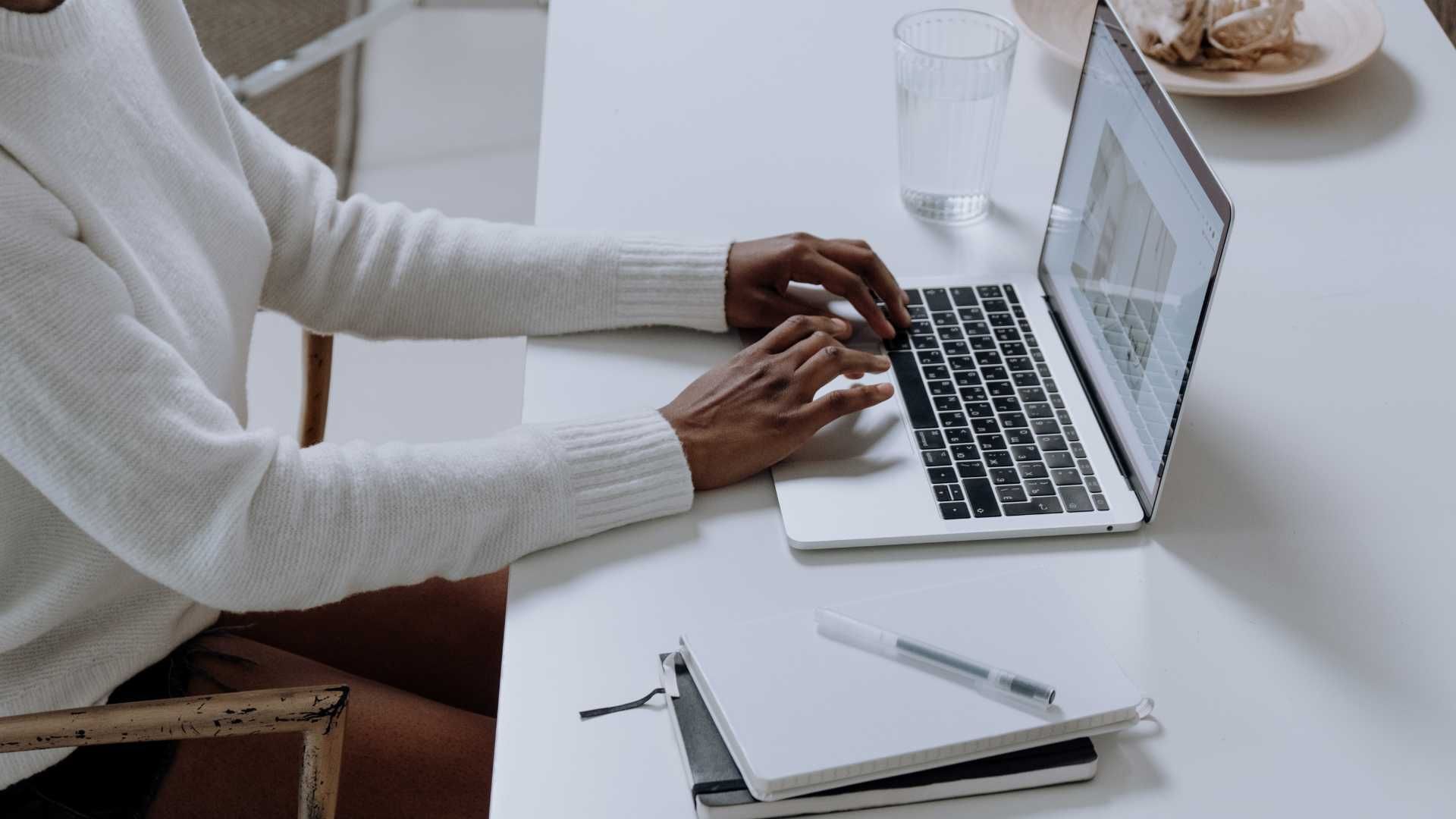 Person typing on laptop at a white desk; a notebook, pen, and glass of water are also present.