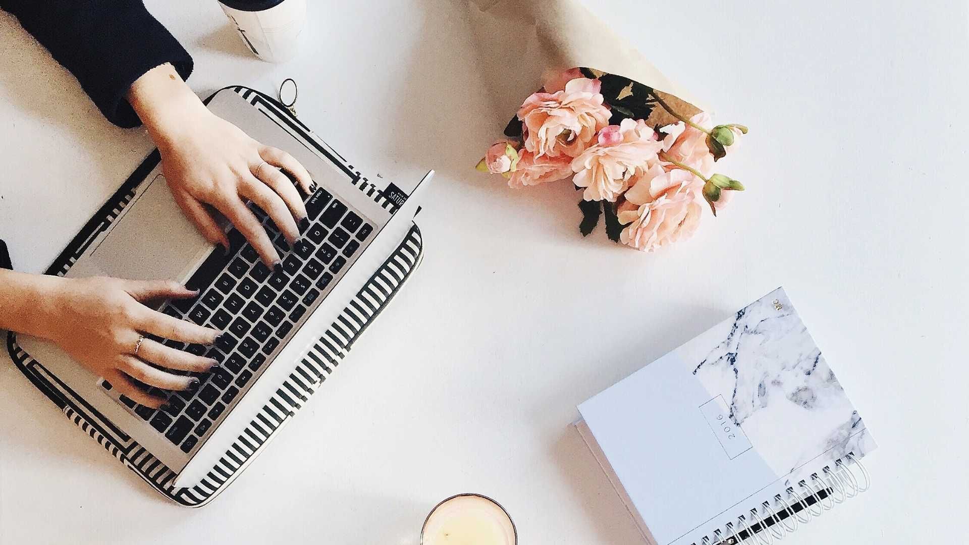 Hands typing on a laptop with a floral bouquet and marble-patterned notebook on a white desk.