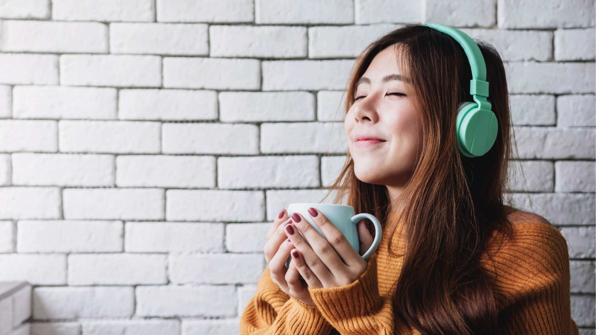 Woman with headphones, holding a mug, eyes closed, smiling, near a brick wall, wearing an orange sweater.
