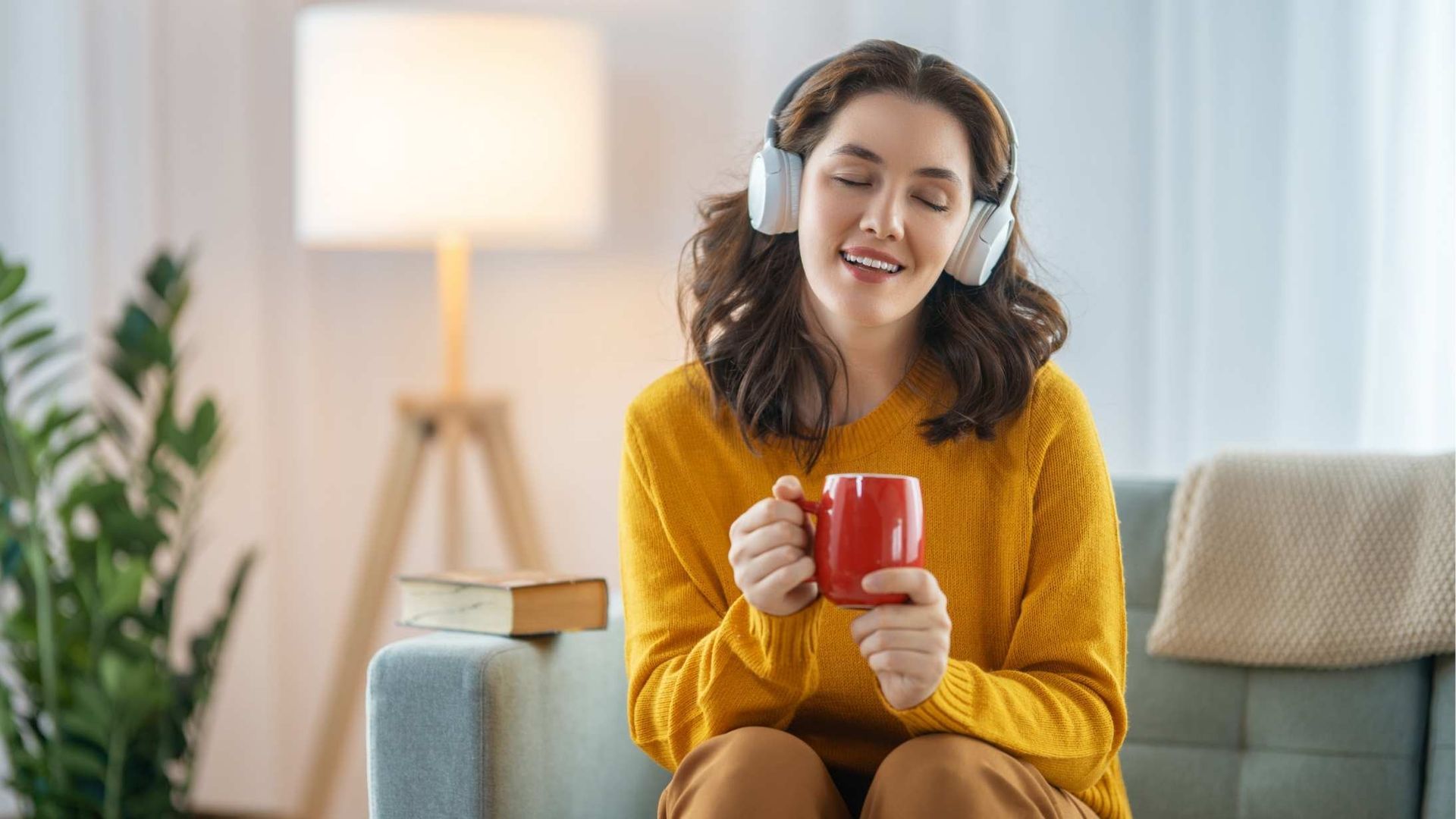 Woman wearing headphones, holding red mug, and smiling. Sitting on a blue couch in a living room.