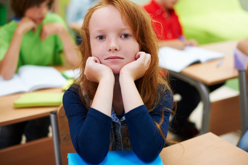 A young girl with red hair is sitting at a desk in a classroom with her hands on her face.