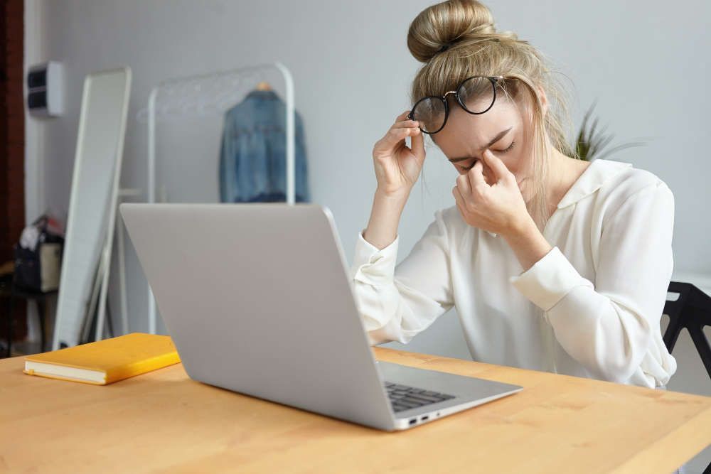 une femme est assise à un bureau avec un ordinateur portable et se frotte les yeux.