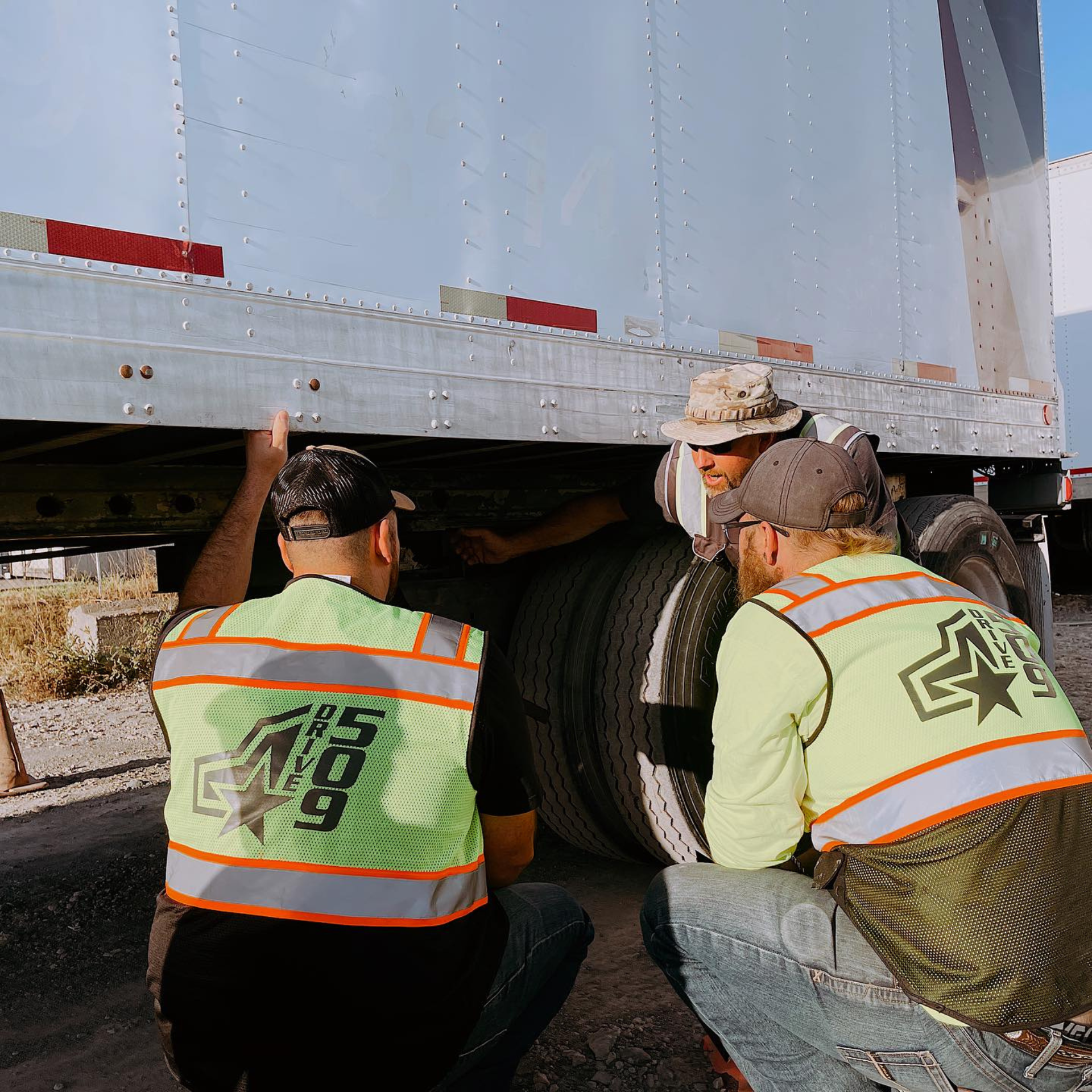 CDL Instructors teaching students how to drive a semi truck kneeled down in the truck yard inspecting the trailer