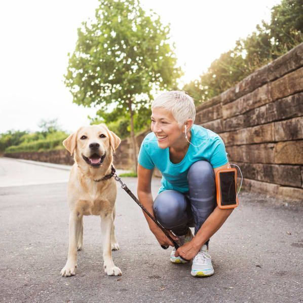 A person with short light hair in a teal athletic shirt crouches on a path to smile at a smiling yellow Labrador retriever.