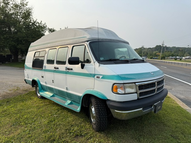 White and teal Dodge camper van parked on grass beside a road under a cloudy sky.