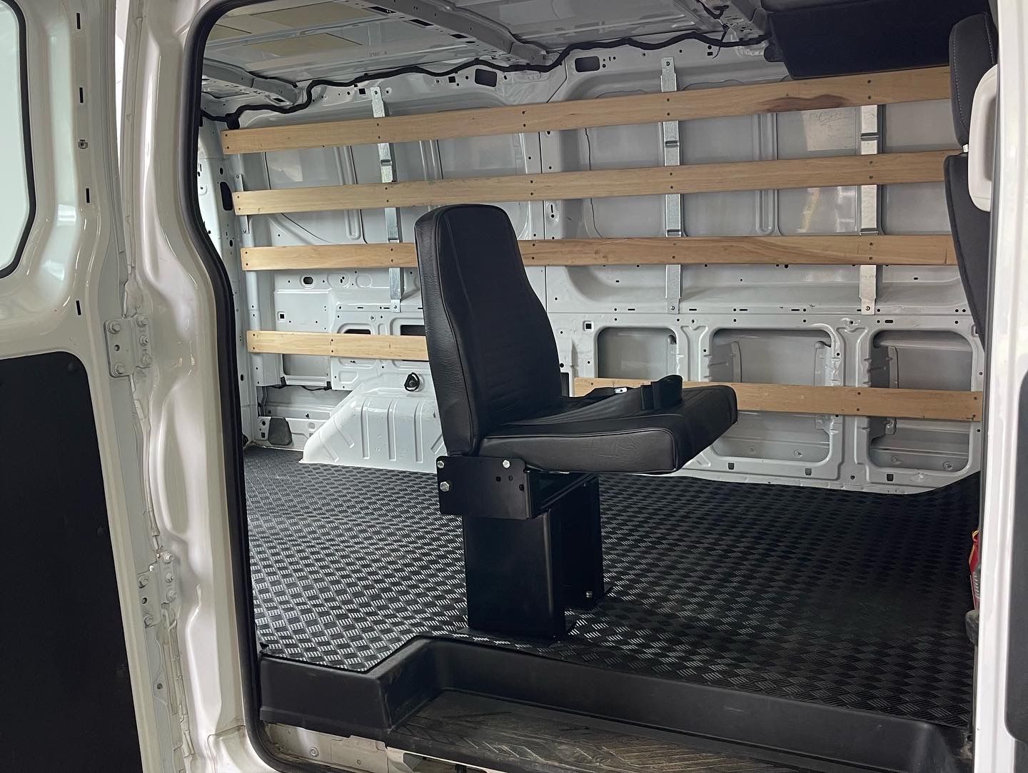 Interior of a white van with a black seat and wooden shelving. Black rubber floor.