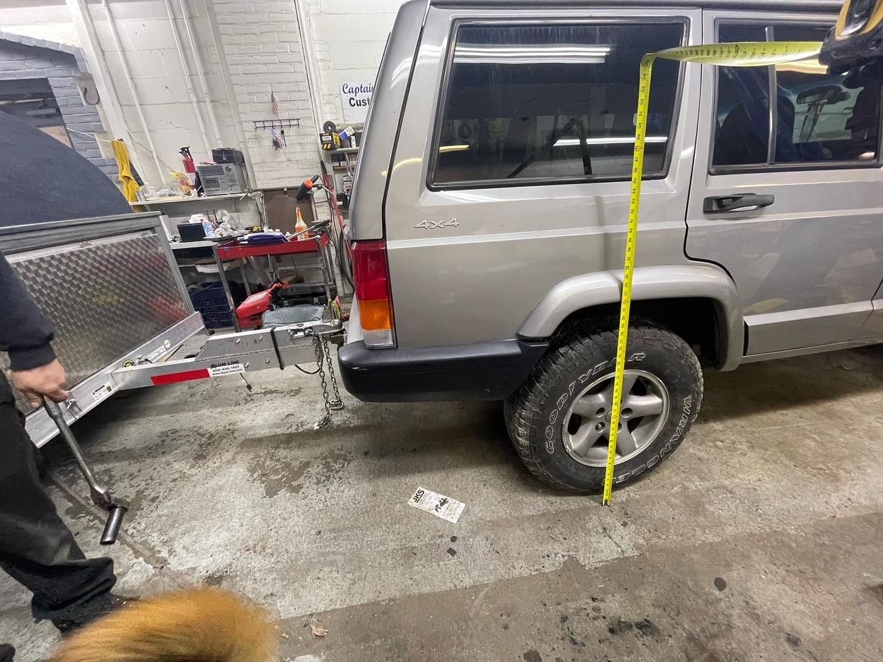 Silver Jeep Cherokee being measured vertically with a yellow tape measure, next to a trailer in a garage.