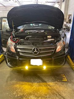 Black Mercedes van with hood open, parked in a repair shop. Front features a brush guard and yellow fog lights.