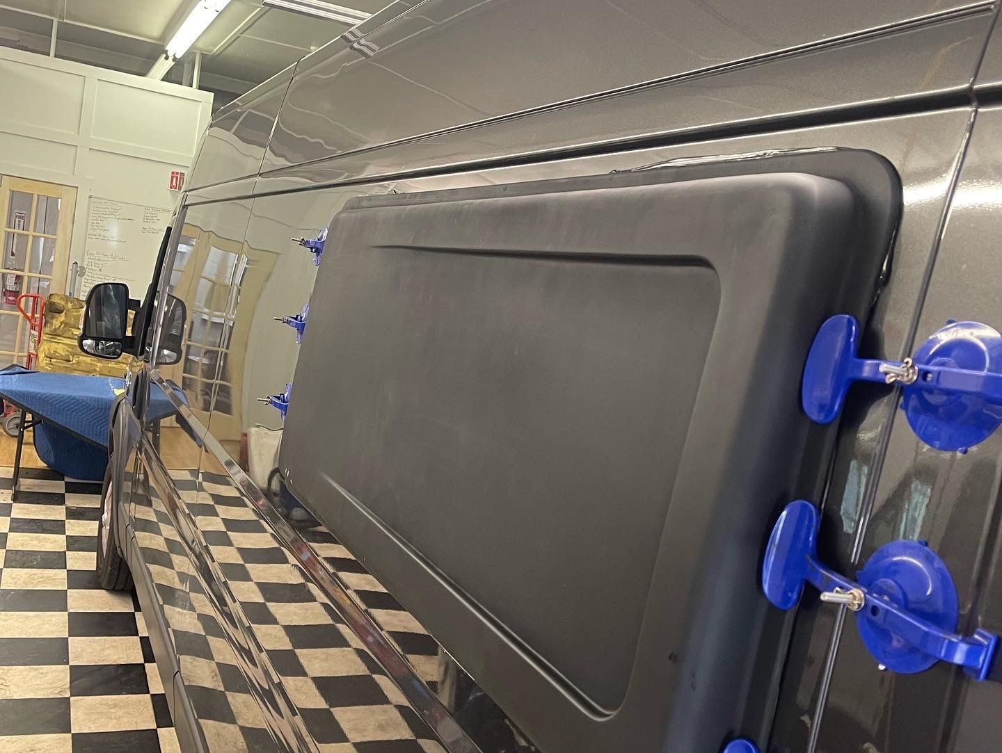 A dark van window is held in place by blue suction cups in a workshop.