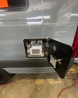 Open fuel door on a gray van reveals water heater components. Black door, red wires visible.