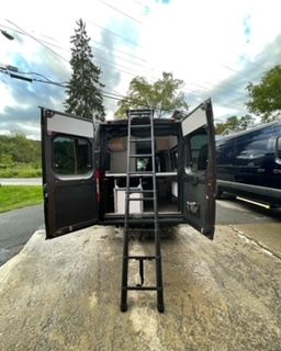 Black van with open doors, interior visible, a ladder extends from it. Outdoors.