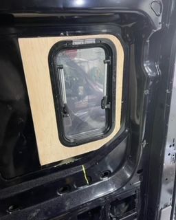Inside a black van, a wooden panel frames a rectangular window with a black gasket, visible from the interior.