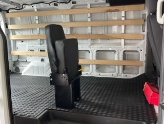 Inside view of a cargo van with a black seat, wooden shelving, and a black rubber floor.