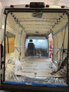 Inside a van's empty interior, insulated with spray foam, plastic sheeting, and wiring visible.