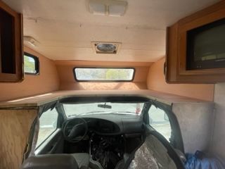 Interior view of a camper van. Dashboard, steering wheel, overhead cabinets, and bunk over the driver's seat.