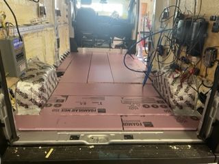 Interior of a van being converted; pink insulation laid on the floor. Electrical wiring and equipment visible on the walls.