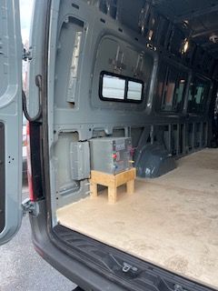 Interior of a van with a battery on a wooden stand. The van is gray with a plywood floor.