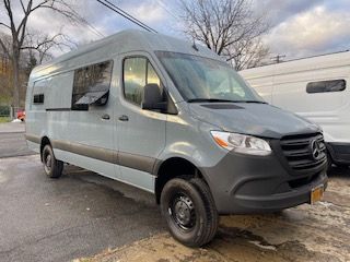 Gray Mercedes Sprinter van parked on pavement. Black tires, side windows, and a black front bumper.
