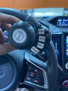 Person holding a custom car steering wheel control with horn, wiper, and washer labels, Subaru interior.