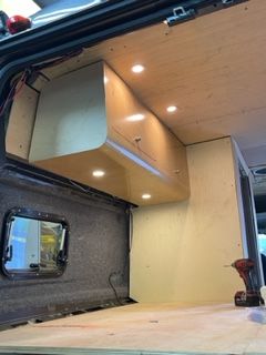 Van interior with built-in cabinetry, spotlights, and window. Beige wood and unfinished surfaces.