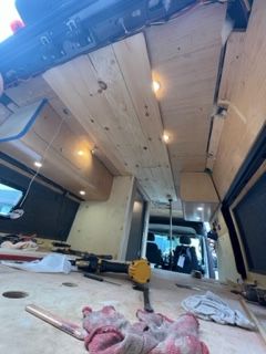 Interior of a van under construction; wooden ceiling and walls with lights. Tools and supplies visible.