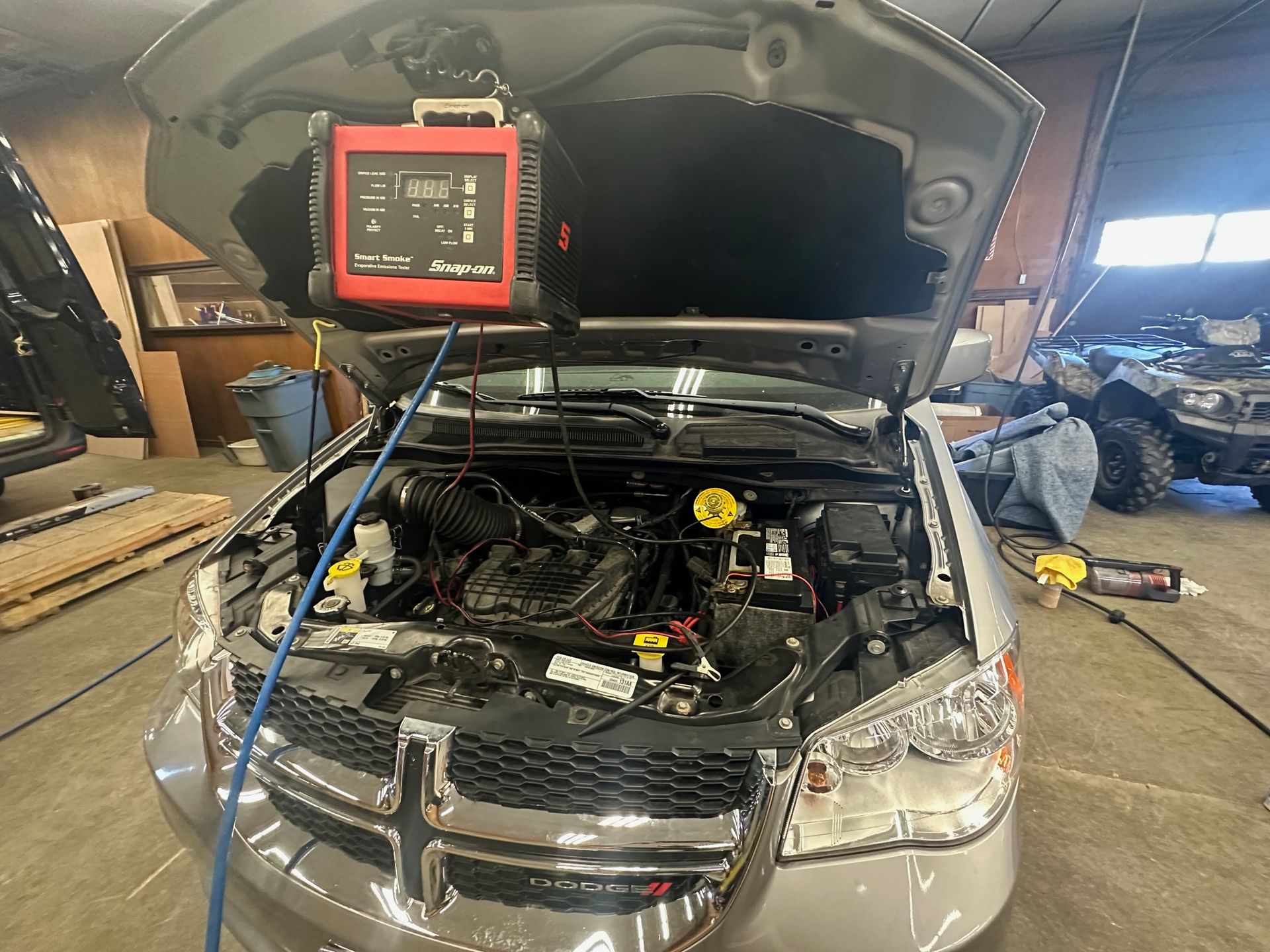 A gray Dodge minivan has its hood open, hooked up to a red battery charger in a garage.