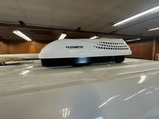 White Dometic air conditioner on the roof of a white van in a garage with fluorescent lights.