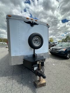 White enclosed trailer with a spare tire, parked in a lot under a cloudy sky.