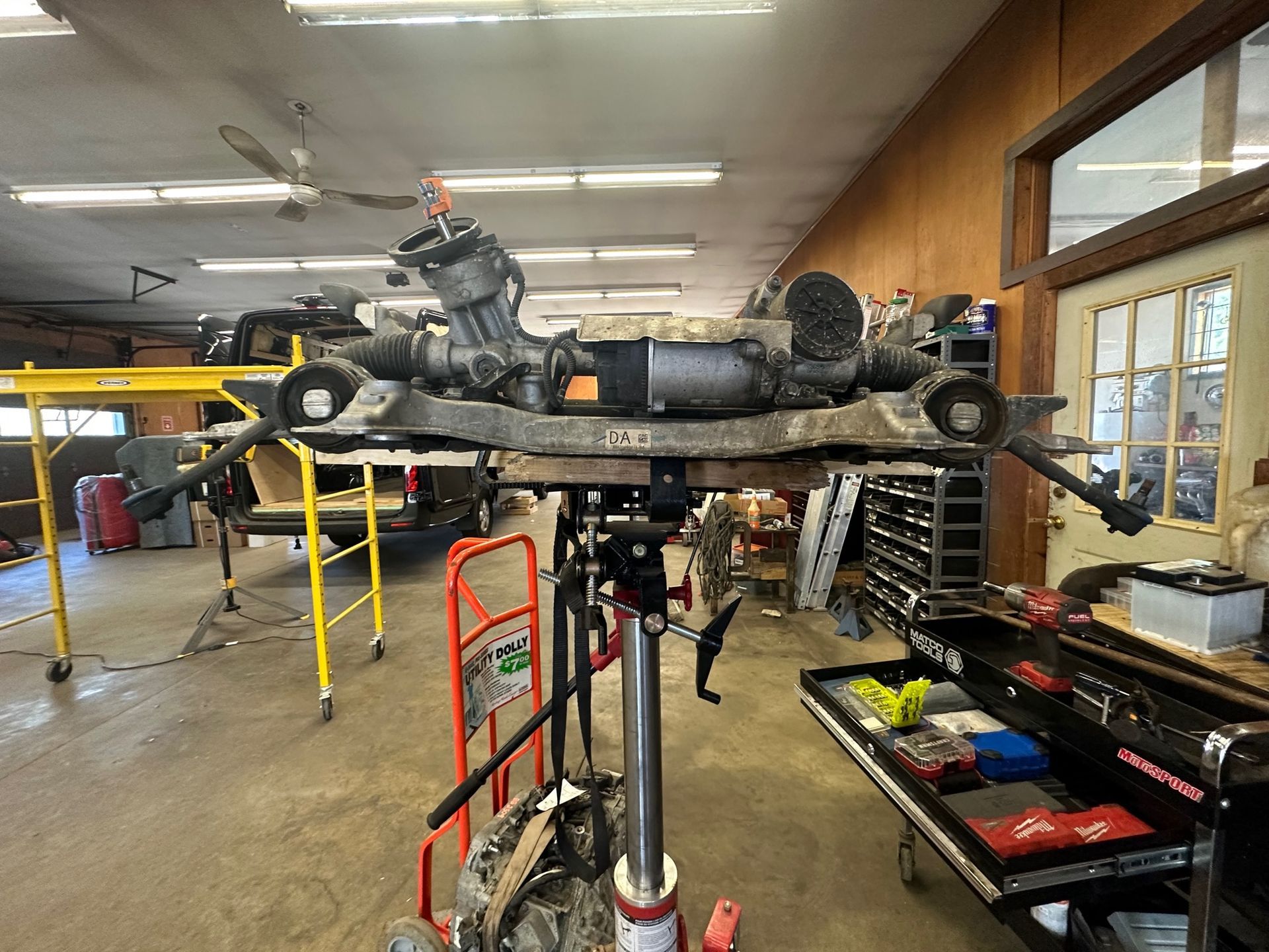 Car's front subframe on a lift in a shop. It's grey with parts and tools around, work in progress.