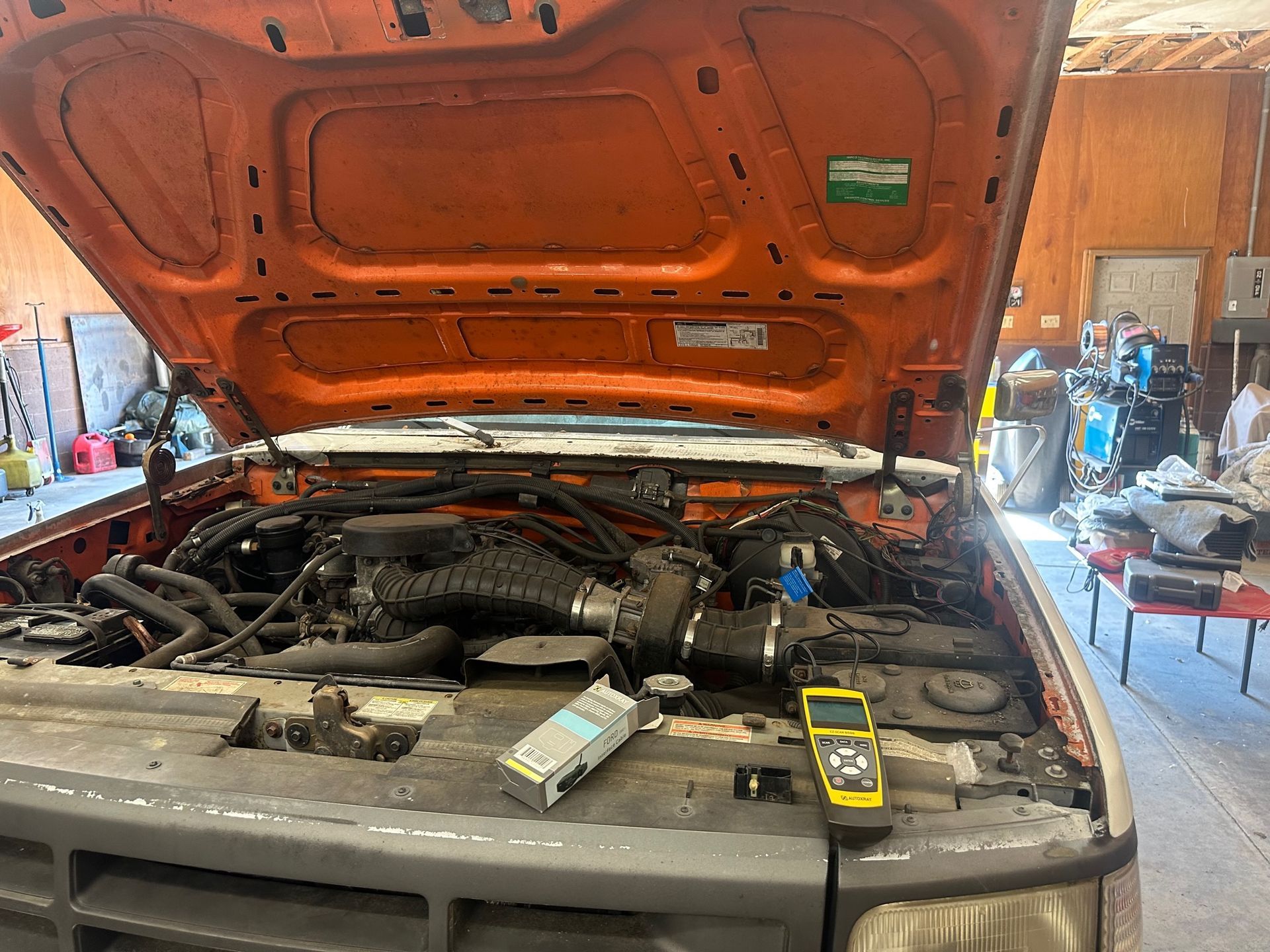 Orange truck hood open, showing engine. Gray vehicle, tools nearby, indoor garage setting.