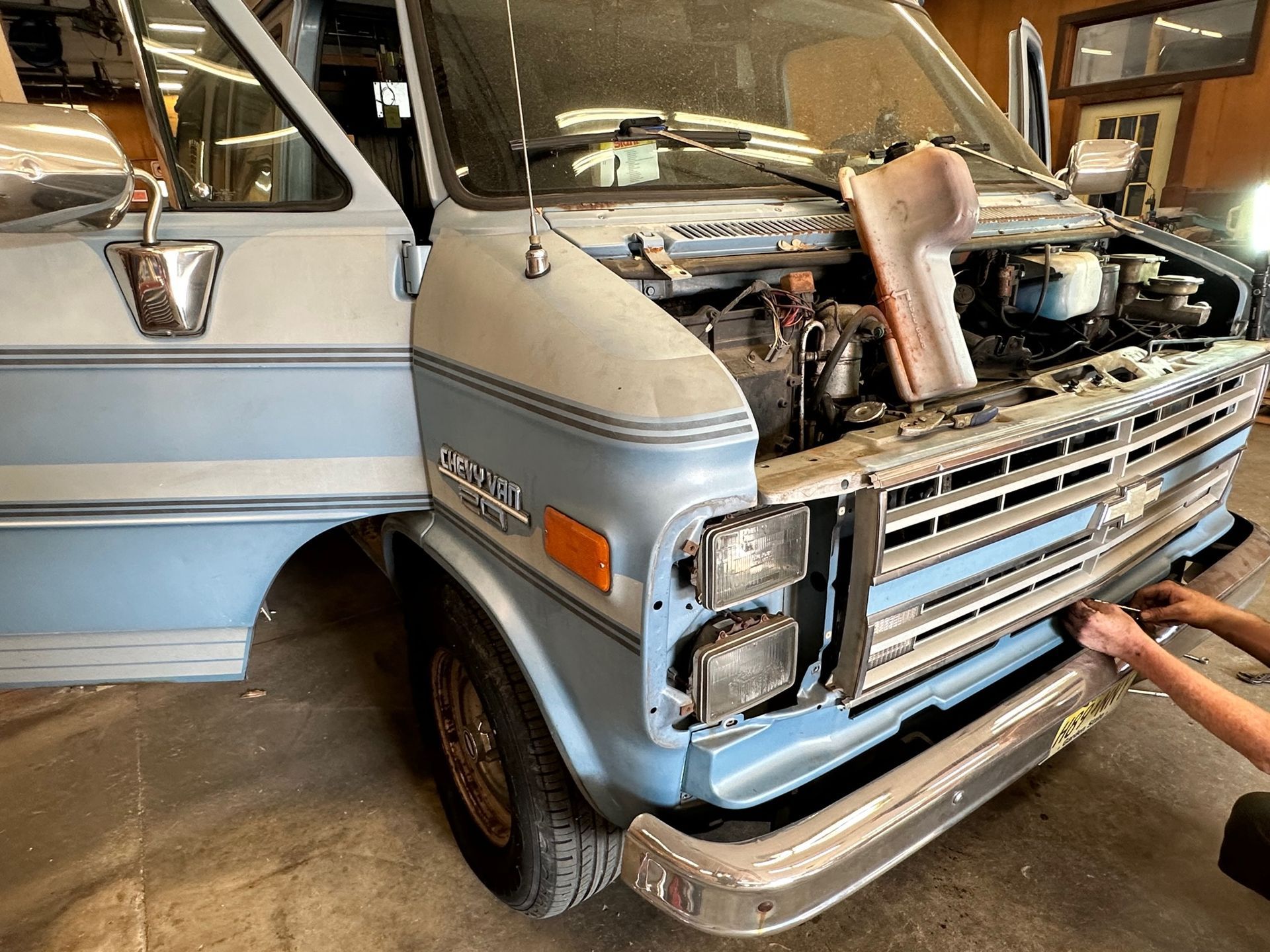 Light blue Chevy van being worked on with open hood. Interior garage setting.