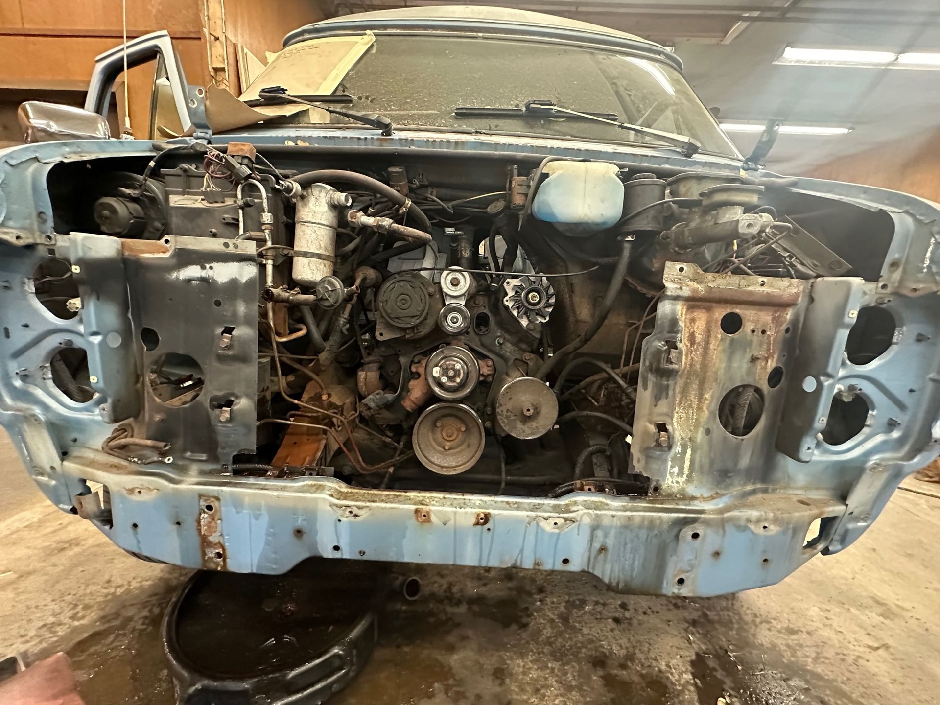 Blue car with the hood open, engine visible, in a garage, appears to be under repair.