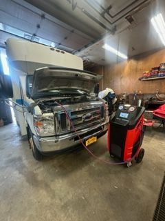 A white RV has its hood open in a garage. An AC machine is connected to the vehicle.