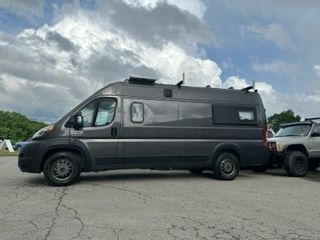 Dark gray camper van parked next to a gray SUV on a paved surface. Cloudy sky.