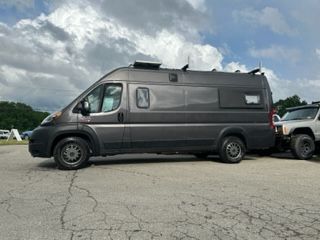 Gray camper van parked next to a white SUV on an asphalt lot under a cloudy sky.
