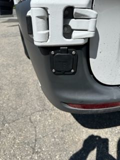 Black trailer hitch connector on a white vehicle, near a hinge. Gray asphalt background.