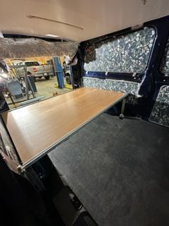 Inside a van: a built-in wooden platform with a gray rug on a blue wall.