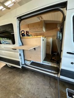 White van interior with wooden cabinets, open sliding door, and fold-down table.