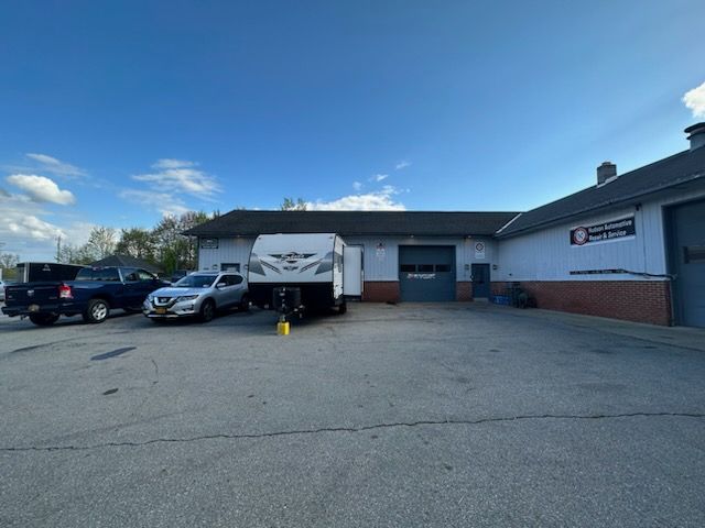A travel trailer is parked outside of an auto repair shop. Cars are parked nearby on a paved lot.