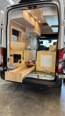 Interior of a van under construction; wooden cabinetry and drawer are visible, with overhead storage.