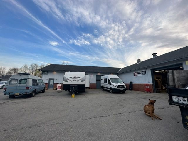 A dog sits on a paved lot in front of a garage. Vans and a camper trailer are parked outside under a blue sky.