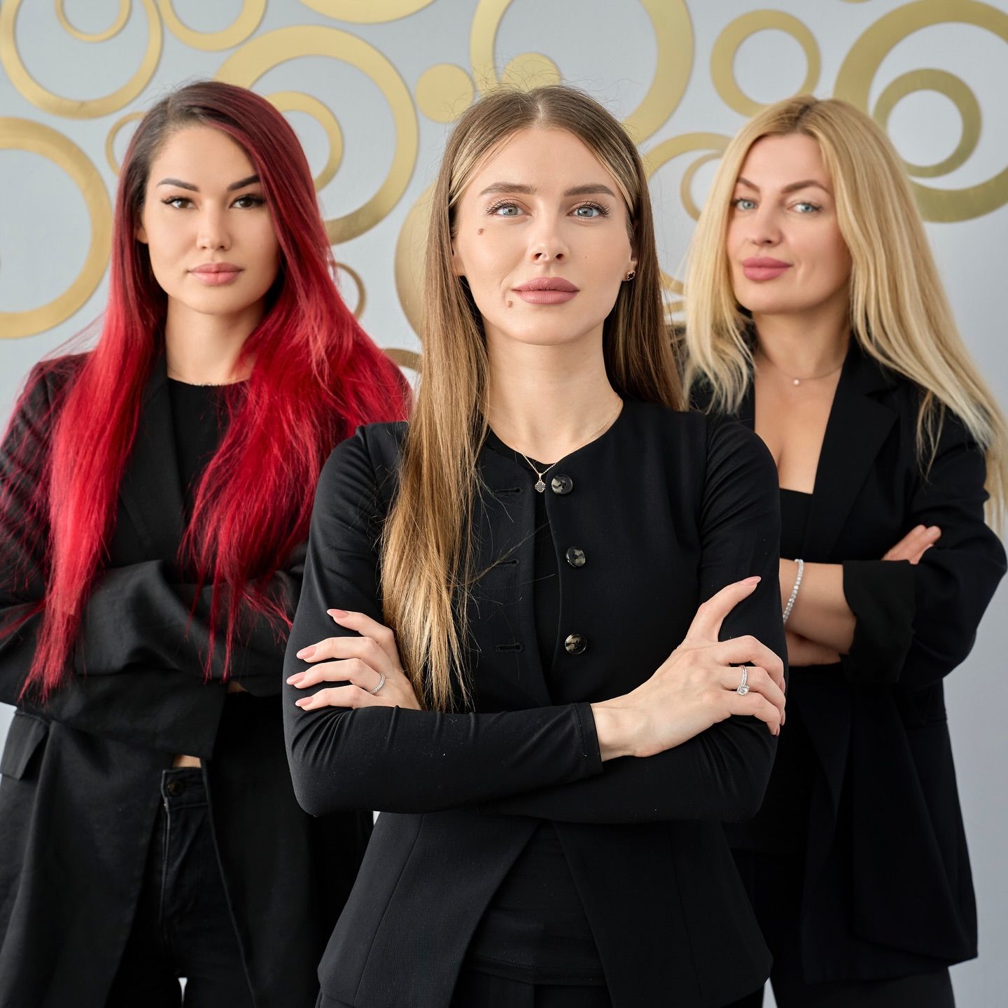 Three women stand with arms crossed in front of a gray wall featuring gold concentric circle designs.