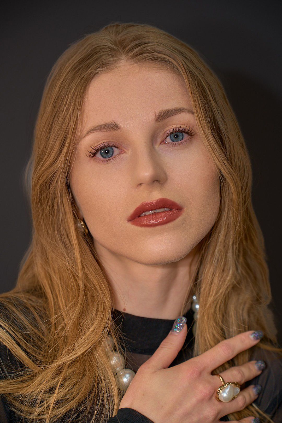 A woman with long blonde hair is wearing a pearl necklace and a ring on her finger.