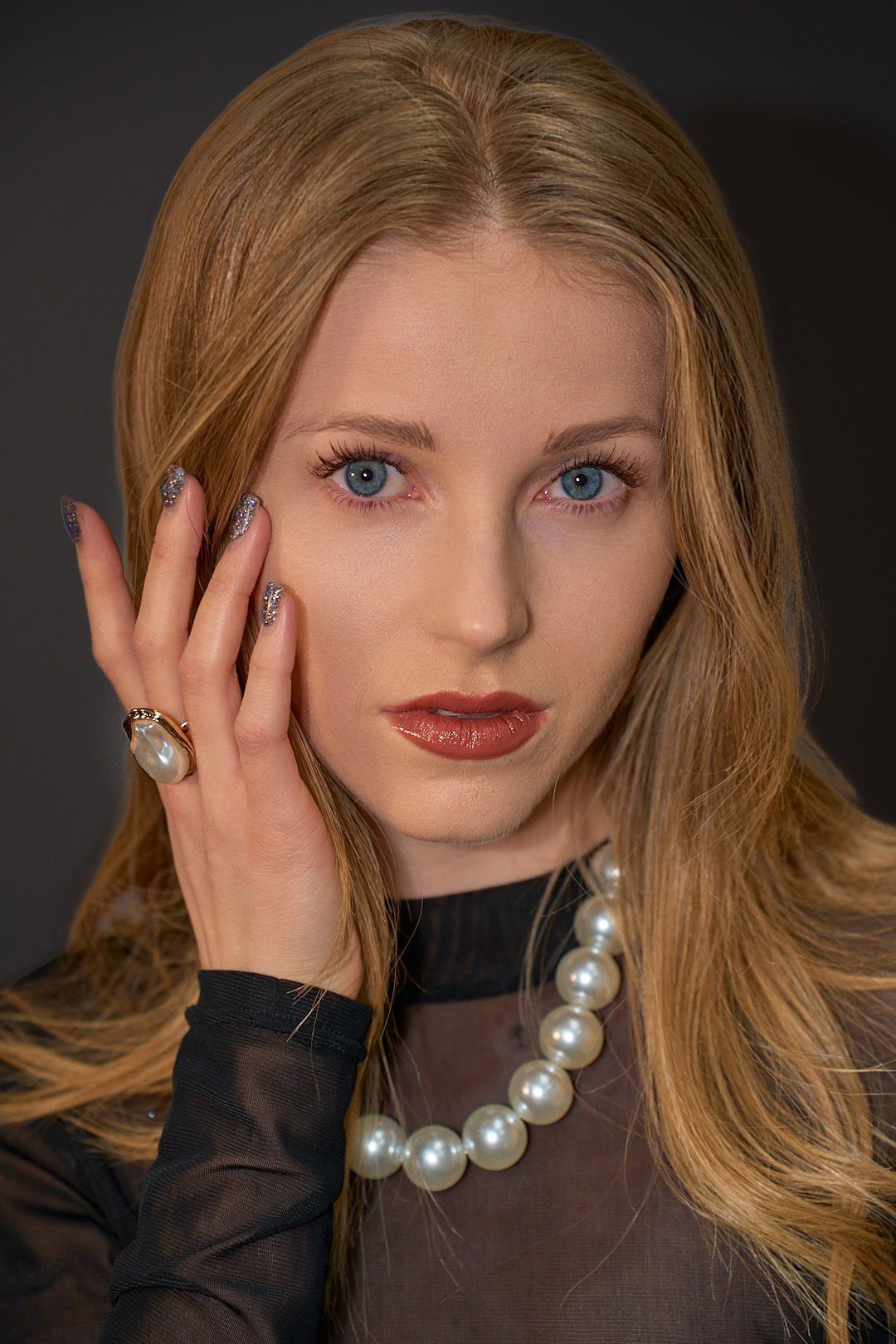 A woman wearing a pearl necklace and a ring is touching her face.