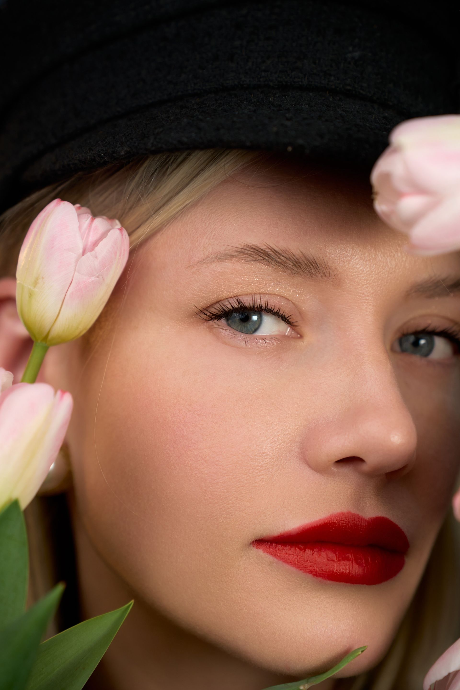 A woman wearing a black hat and red lipstick is holding pink tulips in front of her face.