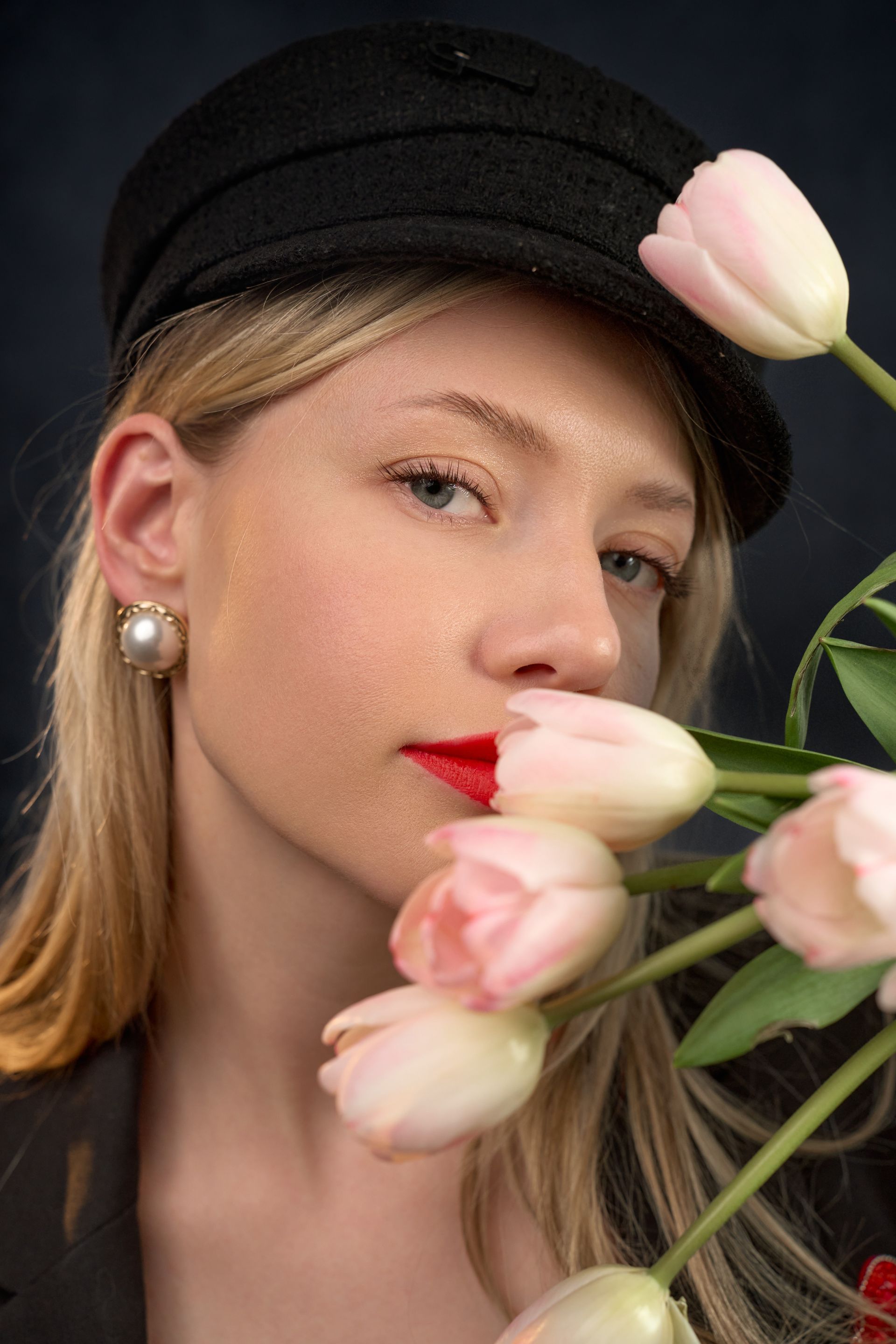 A woman wearing a hat and earrings is holding a bunch of pink flowers in front of her face.
