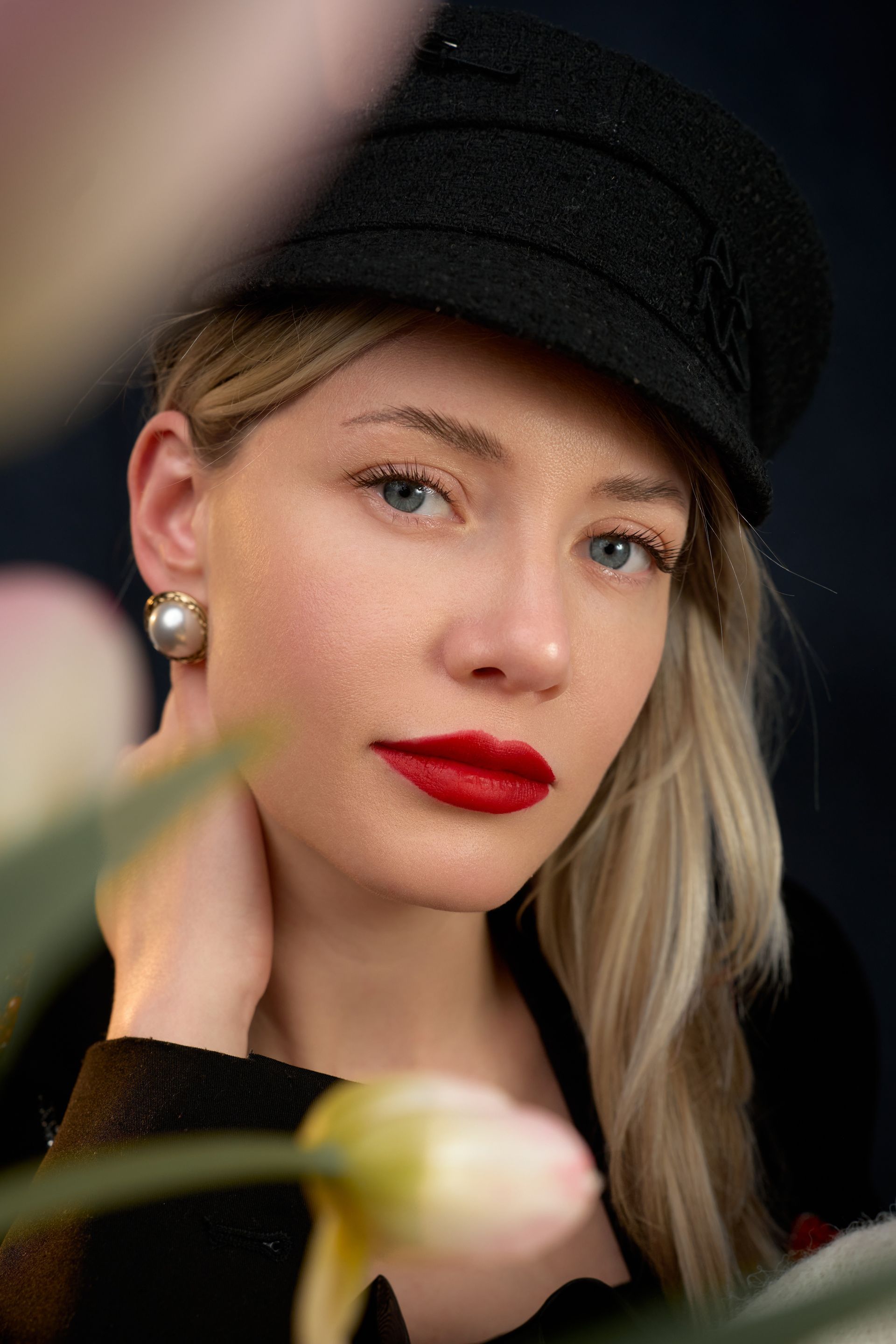 A woman wearing a black hat and red lipstick is standing in front of flowers.
