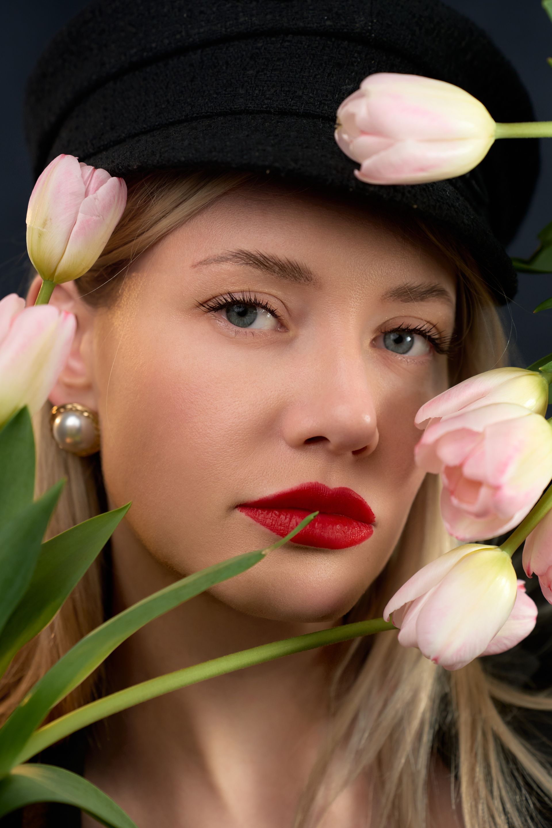 A woman wearing a black hat and red lipstick is holding flowers in front of her face.