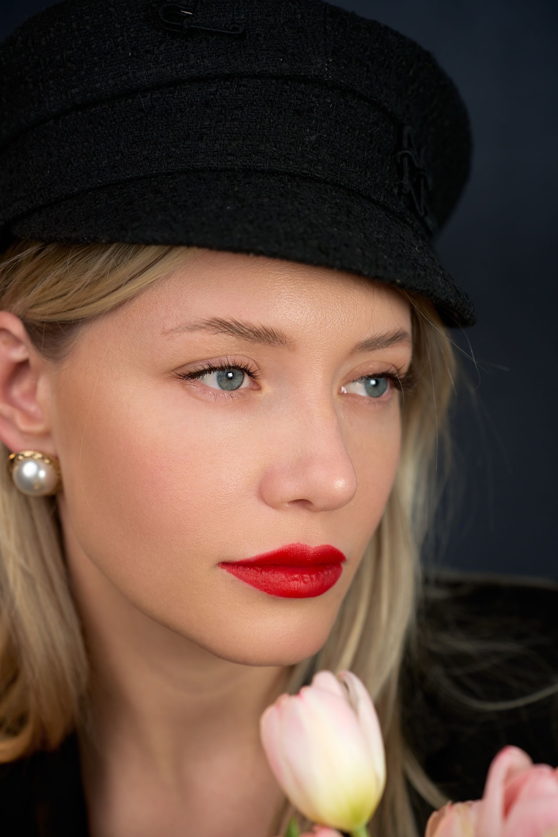 A woman wearing a black hat and red lipstick is holding a pink flower.