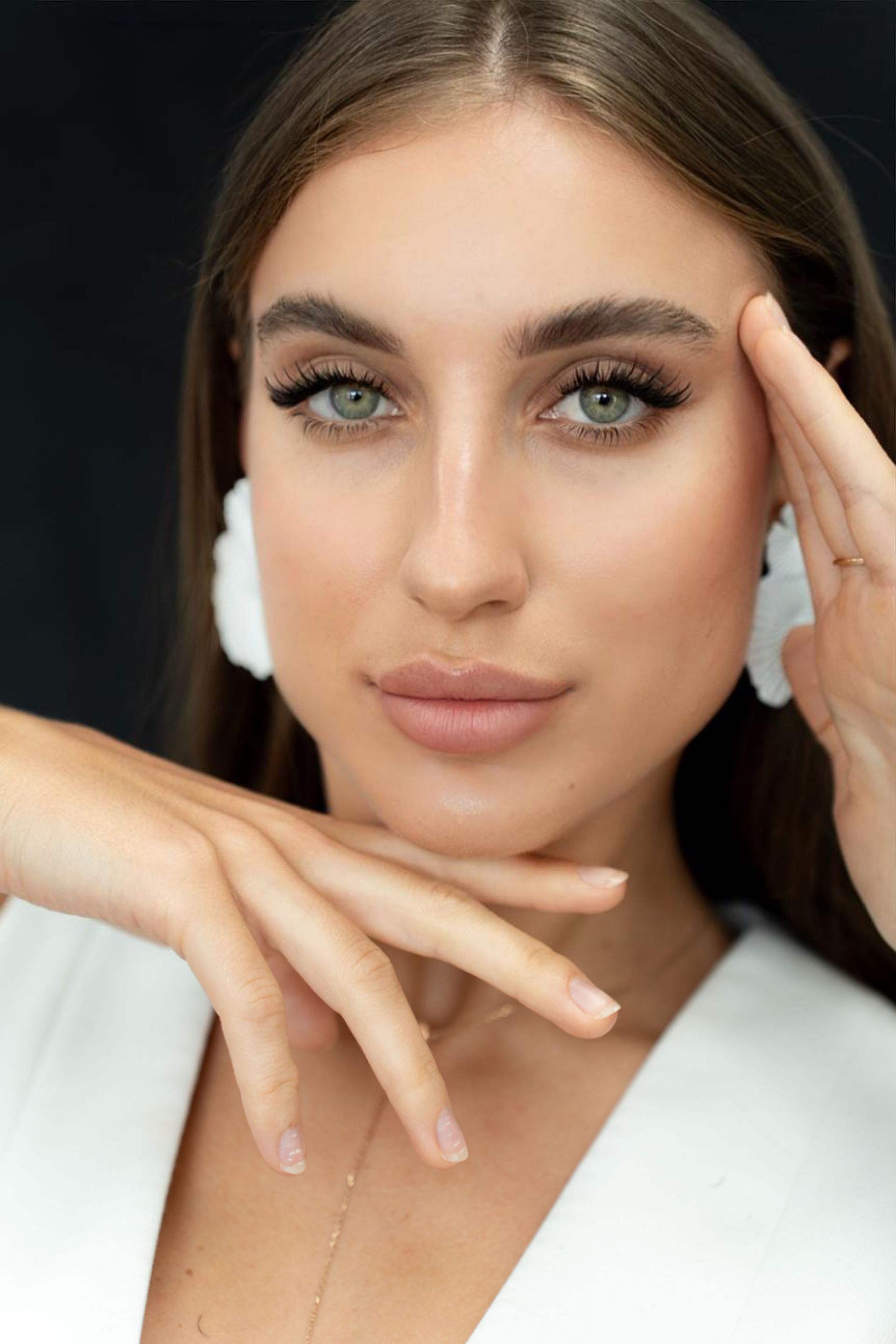 A woman wearing a white jacket and white earrings is touching her face.